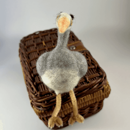A needle-felted great blue heron borb with pipe cleaner legs, facing directly towards the camera, sits on a small wicker case.