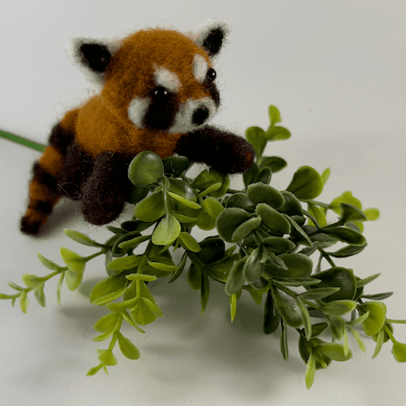 A needle-felted red panda sits facing 3/4 towards the right on top of a plastic branch.
